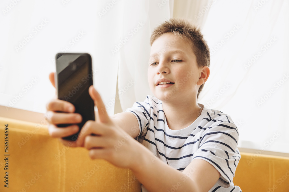 Teenage boy sitting on couch using smartphone texting and having video chat on internet online at home. Child holding mobile phone and looking at screen. Social media. Young blogger influencer Teenage boy sitting on couch using smartphone texting and having video chat on internet online at home. Child holding mobile phone and looking at screen. Social media. Young blogger influencer