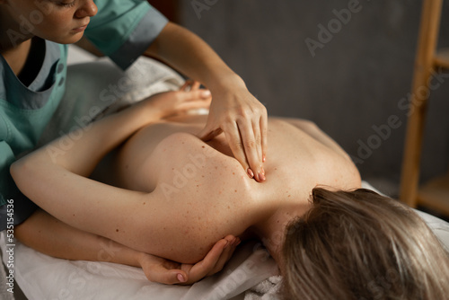 Female masseur massaging back and shoulder blades of young woman lying on a massage table. Professional masseur doing therapeutic massage. Massage spa treatments.