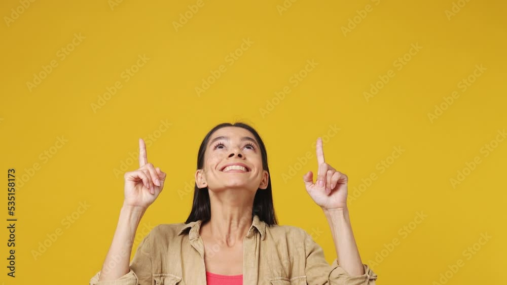 Young promoter fun latin woman wear beige shirt pink t-shirt pointing fingers hands overhead on workspace copy space mockup promo commercial area isolated on plain yellow background studio portrait