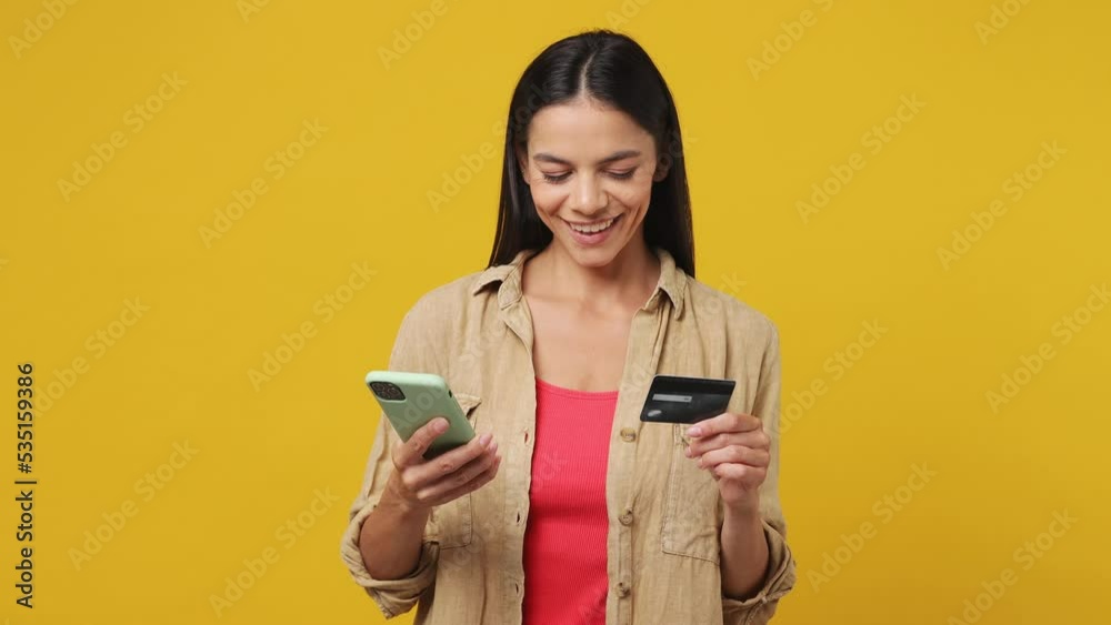 Young latin woman 30s she wear beige shirt pink t-shirt using mobile cell phone hold in hand credit bank card do online shopping order delivery to home booking tour isolated on plain yellow background Young latin woman 30s she wear beige shirt pink t-shirt using mobile cell phone hold in hand credit bank card do online shopping order delivery to home booking tour isolated on plain yellow background