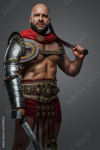 Portrait of ancient gladiator with bare torso posing with dual swords against grey background.