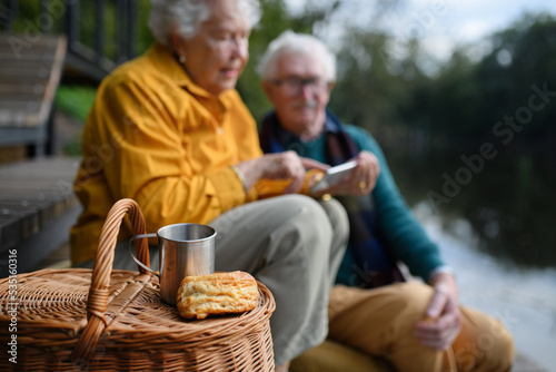 Happy senior couple having picnic and resting near lake after walk.
