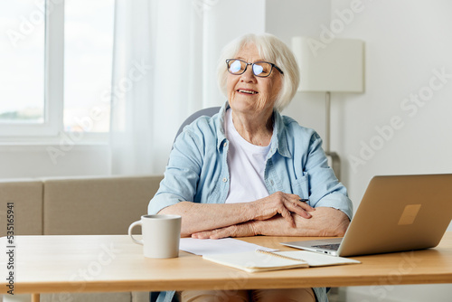 a happy elderly woman with gray hair is sitting at her desk with a laptop and smiling broadly looks away with her hands folded on the table. The concept of working from home