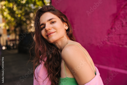 Peaceful young caucasian woman looking at camera smiling, stands outdoor during spring day. Brown hair girl wears casual clothes. Concept solo vacation. © Look!