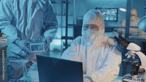 Medium of female Asian scientist in white hazmat suit and respirator sitting at desk in laboratory, using computer, her Black male colleague bringing chemicals and talking