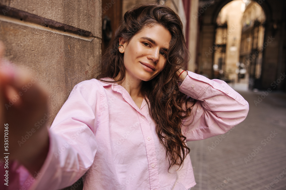 Fototapeta premium Nice young caucasian woman looking at camera taking selfie having fun outdoors. Brown hair girl wears summer light outfit. Concept social life, real emotions.