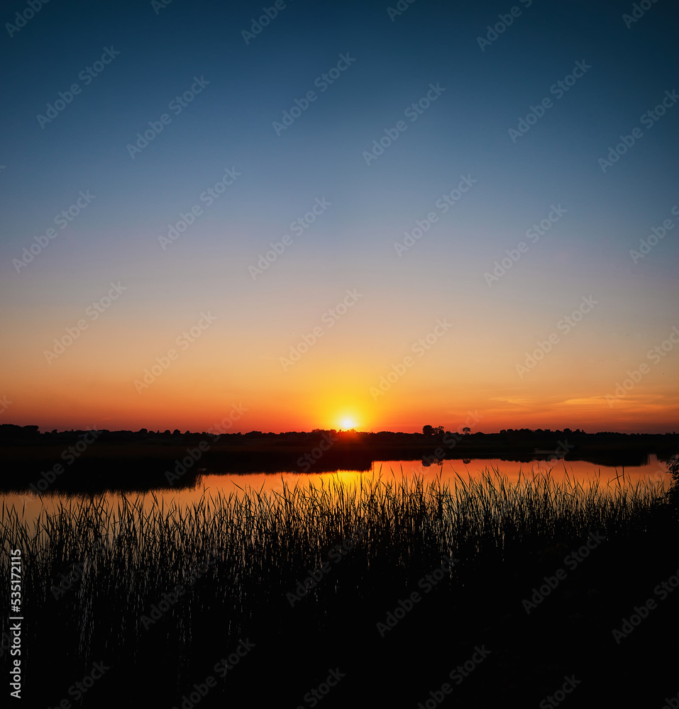 Square photo of a summer sunset over a river with a blue sky in the background and bulrush leaves in the foreground in defocus