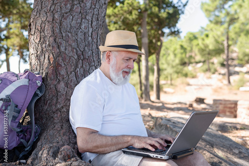 senior digital nomad sitting under the tree using laptop. Active lifestyle concept in retirement