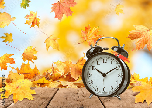 Alarm clock on wooden table and beautiful autumn leaves against blurred background. Time change concept