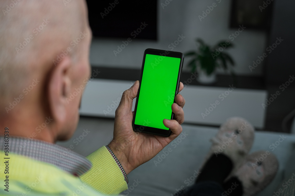 Senior man seated on sofa at home holding vertically mobile device with blank green screen for mockup