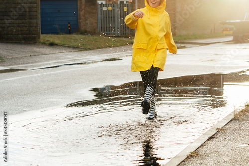 Woman having fun on the street after the rain. Cropped woman wearing rain rubber boots and yellow raincoat walking into puddle with water splash and drops in sunset light. Selective focus