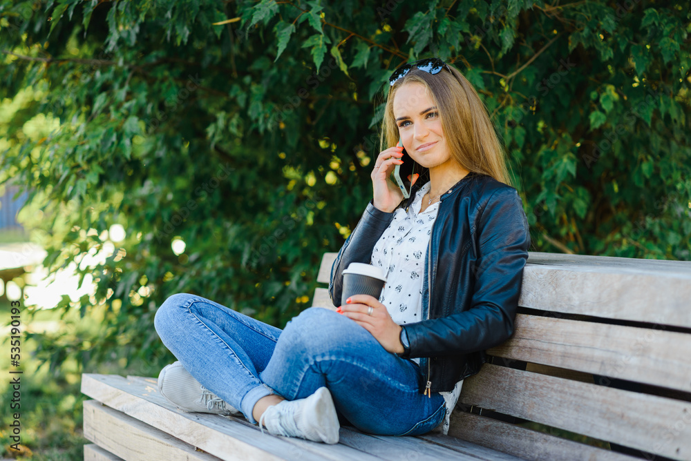 A smiling modern young woman sitting on bench in green park on summer day and talking on smartphone with coffee cup.Girl sitting with a phone and coffe at park.Klaipeda,Lithuania.09-02-2022. A smiling modern young woman sitting on bench in green park on summer day and talking on smartphone with coffee cup.Girl sitting with a phone and coffe at park.Klaipeda,Lithuania.09-02-2022.