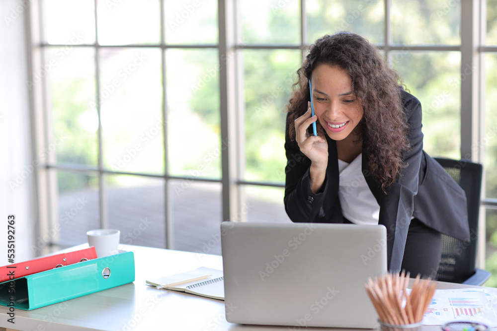 Attractive professional latin female employee worker standing, using laptop computer with paperwork at home workplace. Businesswoman talking on smart mobile phone and communicate with client. Attractive professional latin female employee worker standing, using laptop computer with paperwork at home workplace. Businesswoman talking on smart mobile phone and communicate with client.