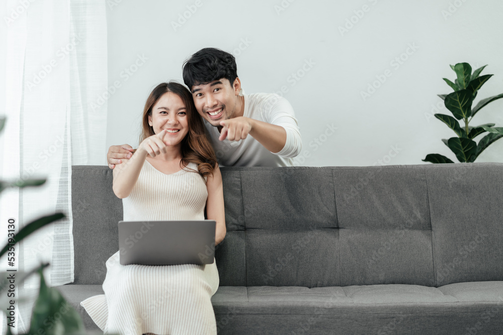 Obraz premium Beautiful pregnant asian couple are sitting on a couch in the living room, looking and pointing at camera with smiles on their faces. Happiness Asian couple use computer laptop planing for the future.