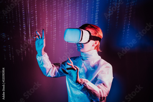 Metaverse concept. Woman in holographic clothes and vr glasses on matrix code background, playing video games with virtual reality headset, trying to touch something with hand in pink blue colors