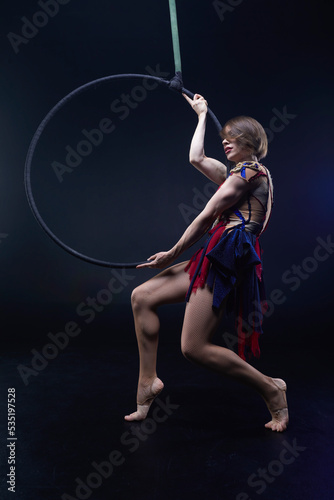 Beautiful woman performing acrobatic element on aerial ring indoors. Circus performer doing trick on arial lira on black background. © Naz