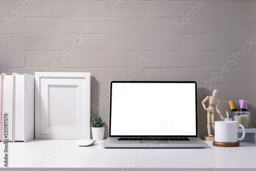 Front view laptop with blank screen, picture frame, books and stationery on white table.