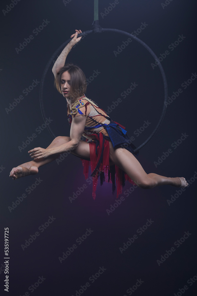 Beautiful woman performing acrobatic element on aerial ring indoors. Circus performer doing trick on arial lira on black background.