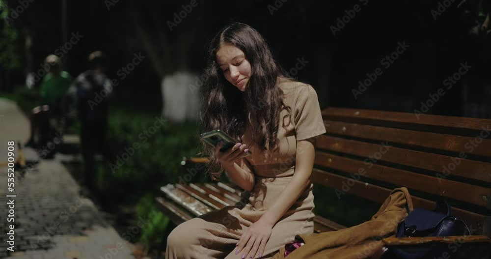 Woman using smartphone sitting on a bench in the night park Woman using smartphone sitting on a bench in the night park