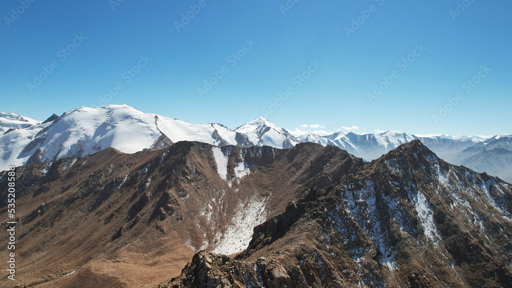 Snowy rocky mountain peaks in a light haze. Aerial view from the drone of the blue sky, light haze and steep cliffs with peaks. A moraine lake can be seen in the distance. Ancient glaciers. Kazakhstan