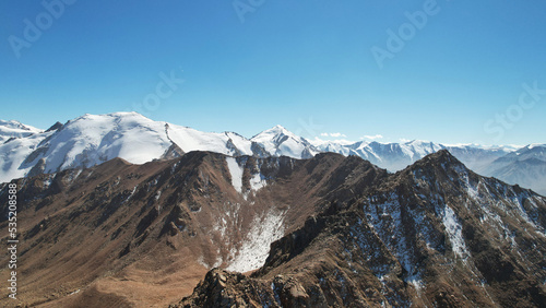 Snowy rocky mountain peaks in a light haze. Aerial view from the drone of the blue sky  light haze and steep cliffs with peaks. A moraine lake can be seen in the distance. Ancient glaciers. Kazakhstan