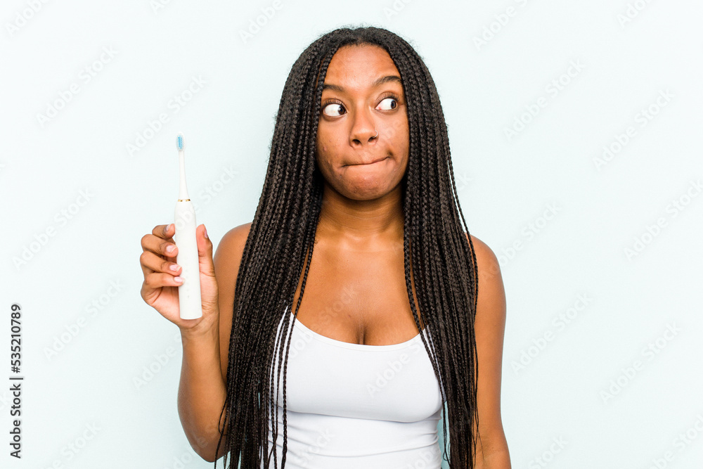 Fototapeta premium Young African American woman holding electric toothbrush isolated on blue background confused, feels doubtful and unsure.
