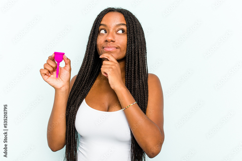 Young African American woman holding menstrual cup isolated on blue background looking sideways with doubtful and skeptical expression. Young African American woman holding menstrual cup isolated on blue background looking sideways with doubtful and skeptical expression.