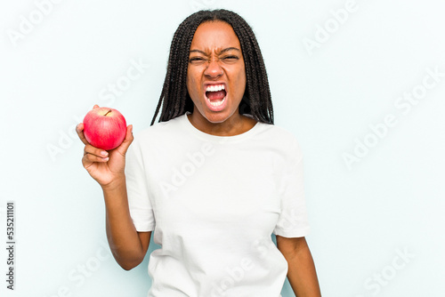 Young African American woman holding an apple isolated on blue background screaming very angry and aggressive.