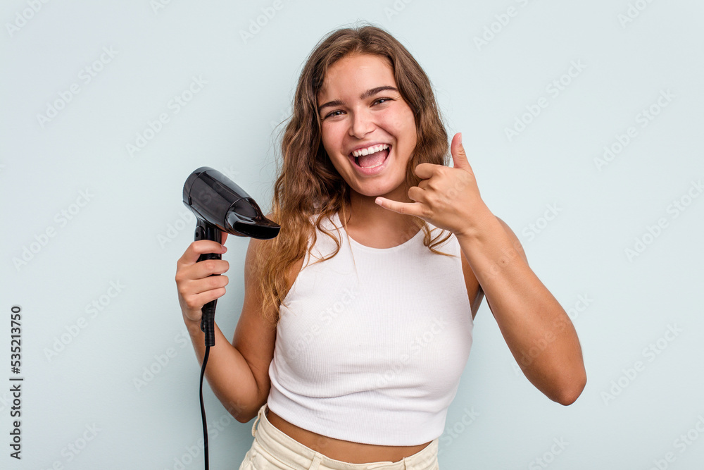 Young caucasian woman holding a hairdryer isolated on blue background showing a mobile phone call gesture with fingers. Young caucasian woman holding a hairdryer isolated on blue background showing a mobile phone call gesture with fingers.