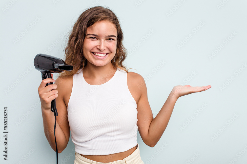 Young caucasian woman holding a hairdryer isolated on blue background showing a copy space on a palm and holding another hand on waist. Young caucasian woman holding a hairdryer isolated on blue background showing a copy space on a palm and holding another hand on waist.