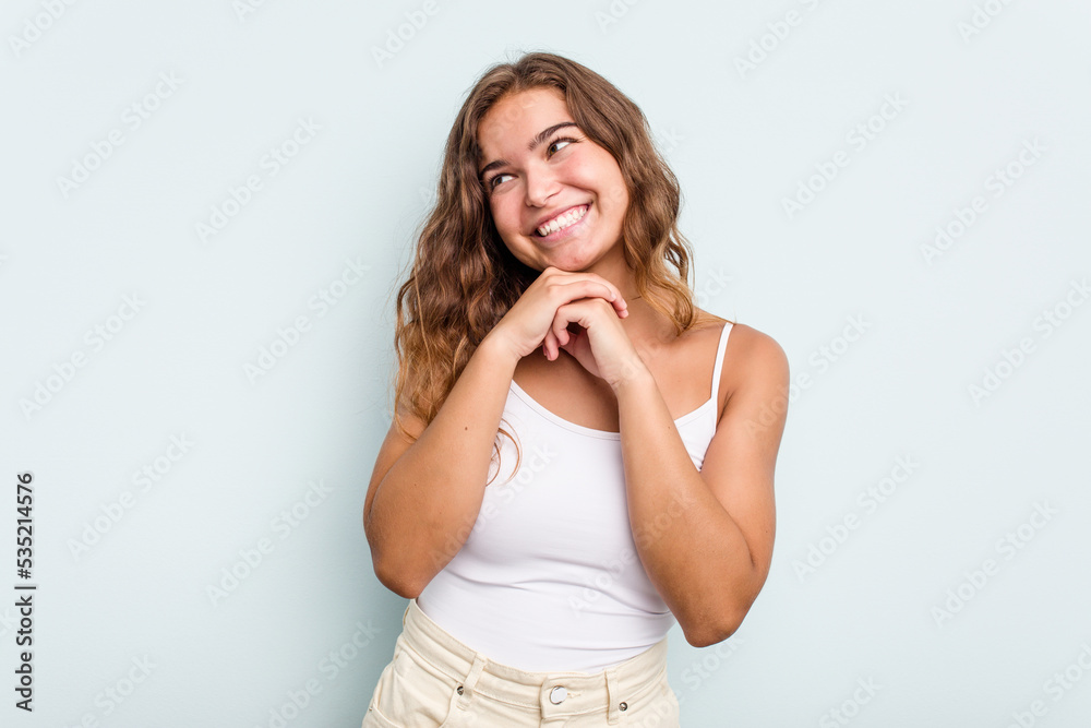 Young caucasian woman isolated on blue background keeps hands under chin, is looking happily aside. Young caucasian woman isolated on blue background keeps hands under chin, is looking happily aside.