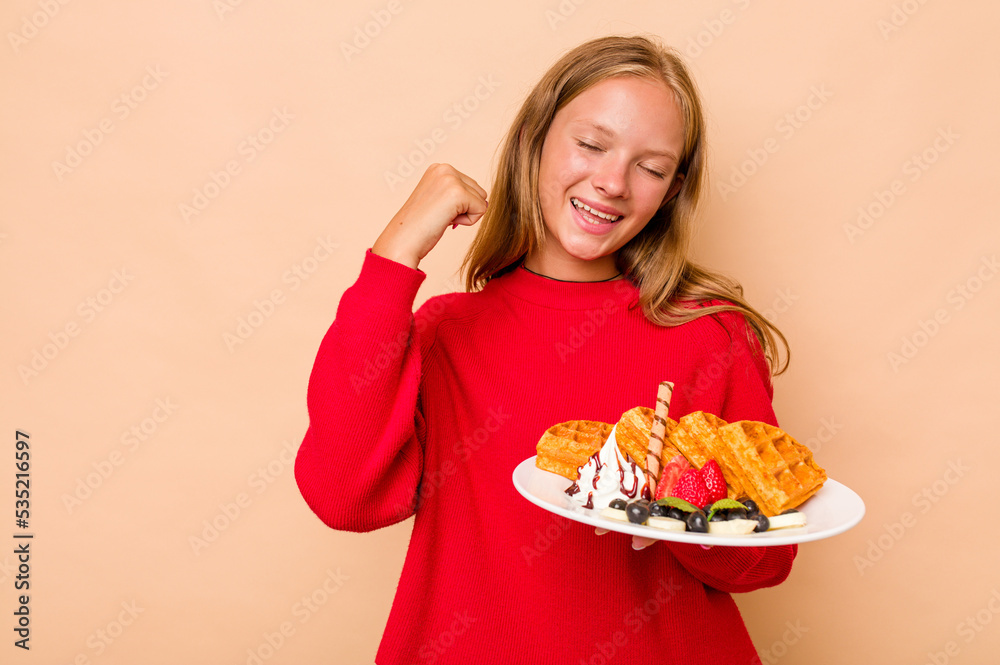 Little caucasian girl holding a waffles isolated on beige background raising fist after a victory, winner concept.