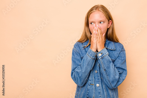 Caucasian teen girl isolated on beige background thoughtful looking to a copy space covering mouth with hand.