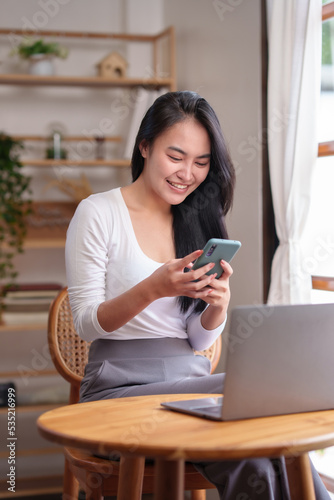 Beautiful Asian woman sitting at home working on her laptop and playing various applications on her smartphone happily and brightly.