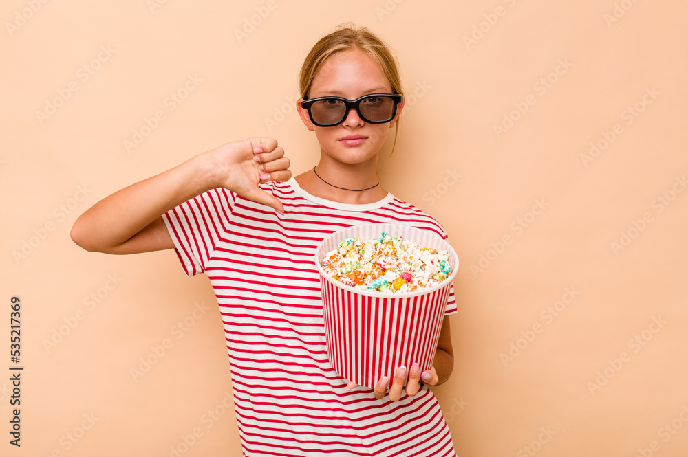 Little caucasian girl eating popcorn isolated on beige background showing a dislike gesture, thumbs down. Disagreement concept.