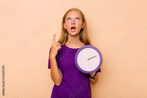 Little caucasian girl holding a clock isolated on beige background pointing upside with opened mouth.