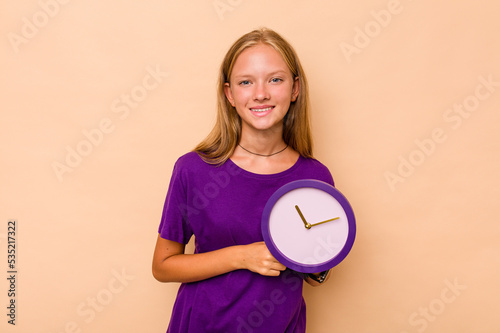 Little caucasian girl holding a clock isolated on beige background laughing and having fun.