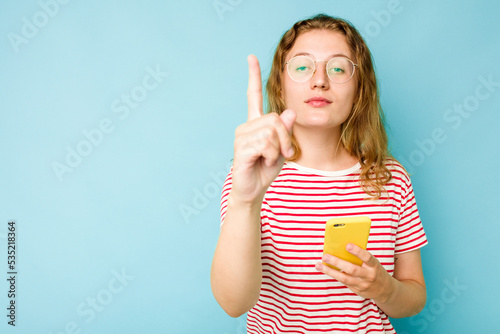 Young caucasian woman holding mobile phone isolated on blue background showing number one with finger.