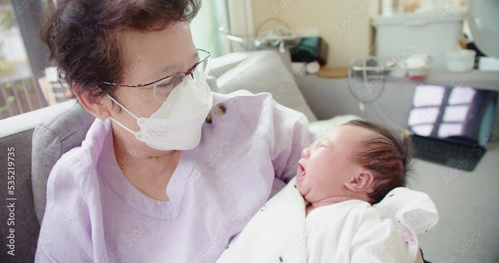 Happy Asian grandmother with protective mask talking looking at granddaughter newborn baby infant in grandmother comfortable arms at home, retired grandparent take care of grandchild, childhood Happy Asian grandmother with protective mask talking looking at granddaughter newborn baby infant in grandmother comfortable arms at home, retired grandparent take care of grandchild, childhood