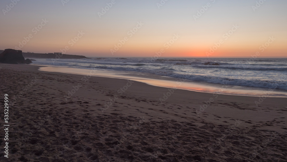 Sunset view of sand beach Praia Grande de Almograve with ocean waves in pink and red blue hour light, clear blue sky. Rota Vicentina coast, Almograve, Portugal
