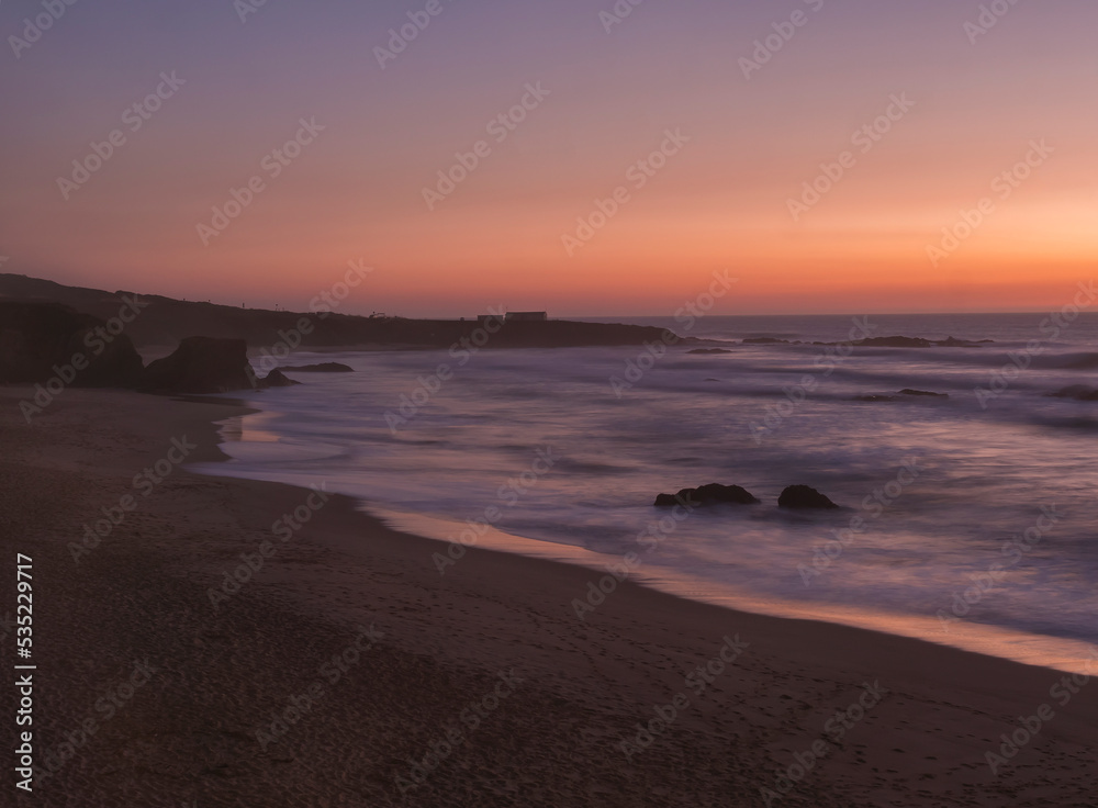 Mysterious long exposure view of sand beach Praia Grande de Almograve with blurred ocean waves in pink orange and purple light. Blue hour after sunset at Rota Vicentina coast, Almograve, Portugal