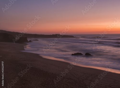 Mysterious long exposure view of sand beach Praia Grande de Almograve with blurred ocean waves in pink orange and purple light. Blue hour after sunset at Rota Vicentina coast, Almograve, Portugal