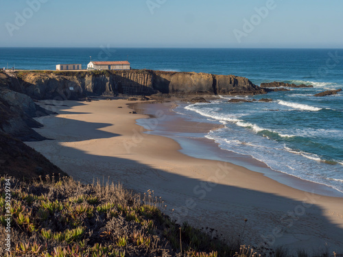 view of Praia Grande de Almograve sand beach with ocean waves, cliff, green vegetation and small beach house. Clear blue sky. Rota Vicentina coast, Almograve, Portugal photo