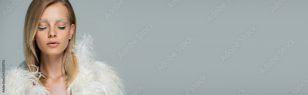 young woman with winter makeup posing with closed eyes in stylish faux fur jacket with white feathers isolated on grey, banner.