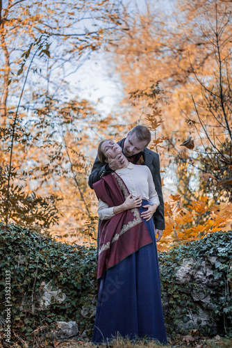 Vampire man wearing victorian suit in the autumn forest bites a woman. Vintage medieval concept style.