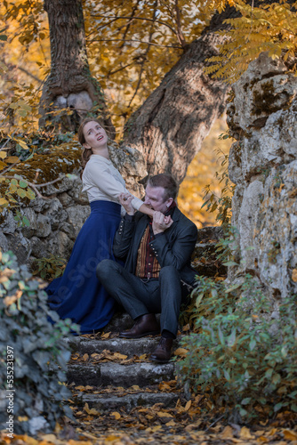 Vampire man wearing victorian suit in the autumn forest bites a woman. Vintage medieval concept style.