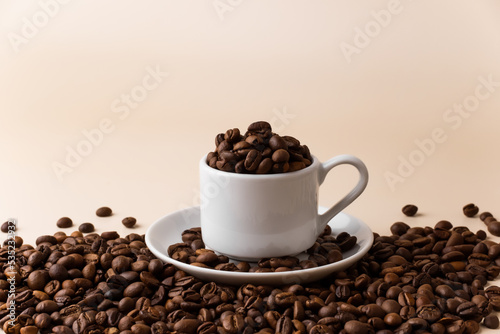 Espresso cup filled with coffee beans surrounded by a large number of coffee beans on a light beige background close-up.