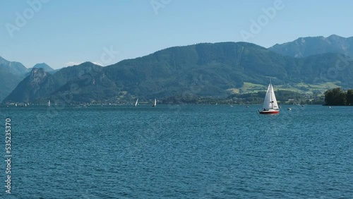 Wide cinematic panorama landscape natural shot with sailing boat on a lake river with clean clear blue water on gorgeous green forest mountains background. Static camera view of a summer vacation day (ID: 535235370)