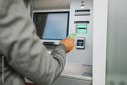 Handsome middle age businessman with eyeglasses standing on city street and using ATM machine to withdraw money from credit or debit card. Hand close up.