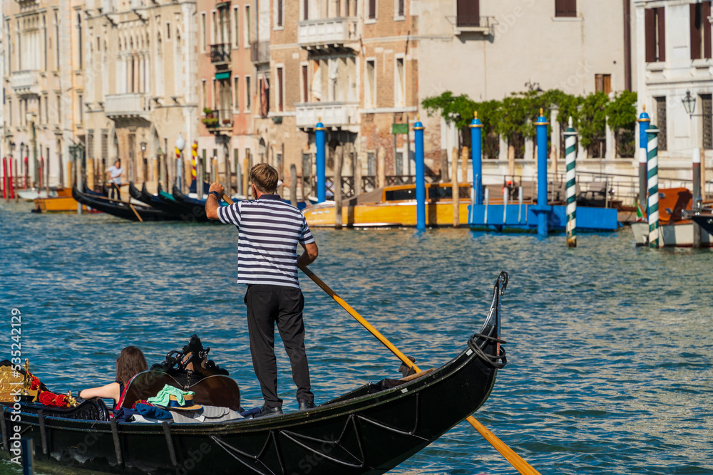 A gondolier standing in a gondola controls the boat with an oar in his hands, a woman floats along the canal in a gondola along the coast of the street of the city of Venice, Venetian gondolier